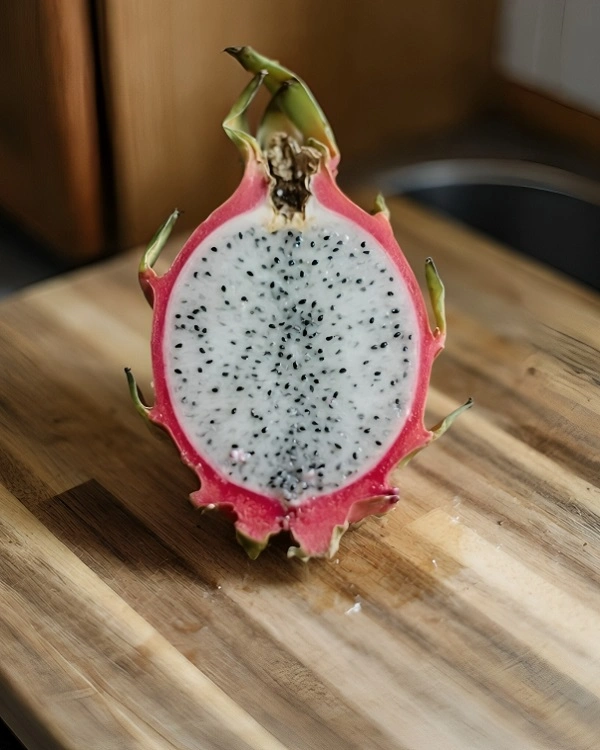 A freshly sliced pink dragon fruit pitaya with white flesh and black seeds on a wooden cutting board.