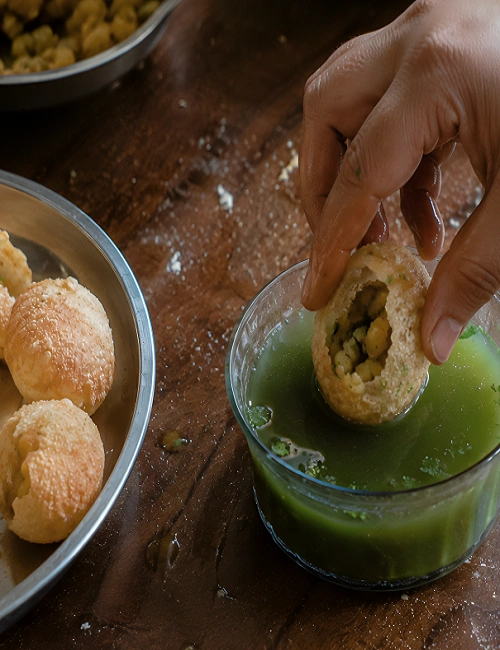A hand dipping a stuffed puri into a bowl of chilled green mint pani.