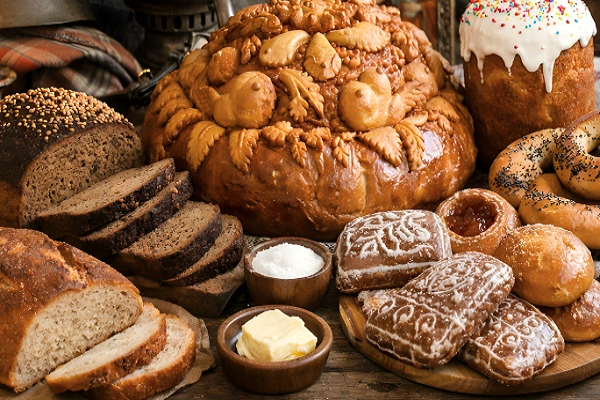 A rustic table displaying an assortment of traditional Russian breads including dark rye Borodinsky, decorative karavai, glazed Easter kulich, ring-shaped bubliki with poppy seeds, and honey pryaniki, surrounded by vintage cultural items and warm lighting.