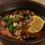 Solyanka soup in a rustic bowl with mixed meats, olives, lemon slice, and fresh herbs