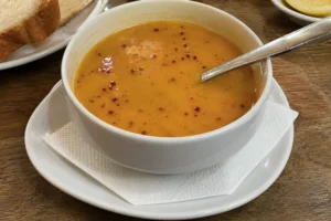 Mercimek çorbası (Turkish red lentil soup) served in a white bowl with bread and chili flakes on a wooden table