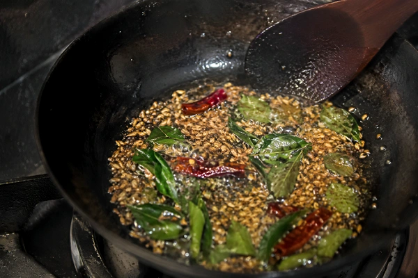 Whole spices blooming in hot oil with curry leaves, cumin seeds, and dried chilies in a pan