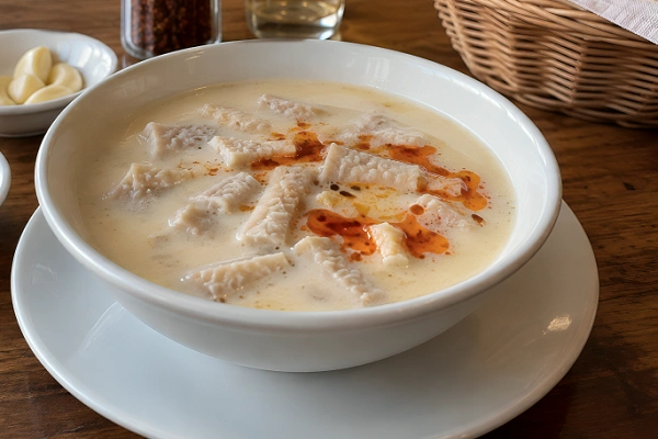 White porcelain bowl of offal soup topped with red pepper oil, served next to raw garlic cloves and a bread basket at a Turkish eatery.