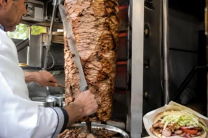 döner kebab meat being sliced from vertical rotisserie in a street food stall with fresh wrap and vegetables