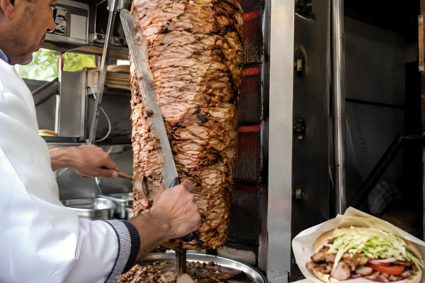 döner kebab meat being sliced from vertical rotisserie in a street food stall with fresh wrap and vegetables