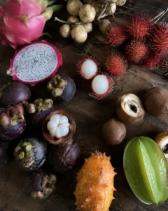 A colorful assortment of fresh exotic tropical fruits including dragon fruit, mangosteen, rambutan, and star fruit on a dark wooden table.