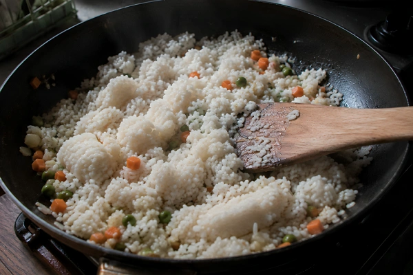 Adding chilled white rice over cooked vegetables in a pan.