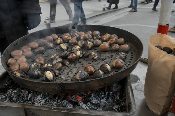 A Turkish street vendor preparing hot chestnuts over burning coals with paper bags ready on the cart.