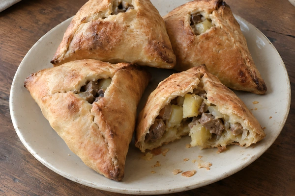 Freshly baked golden brown echpochmak meat pies served on a round plate, with one pie cut open showing the potato and beef filling.