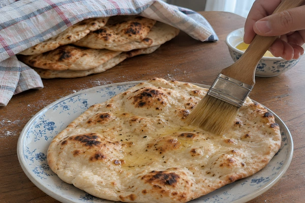 Pastry brush spreading melted butter over a warm fresh naan on a patterned plate
