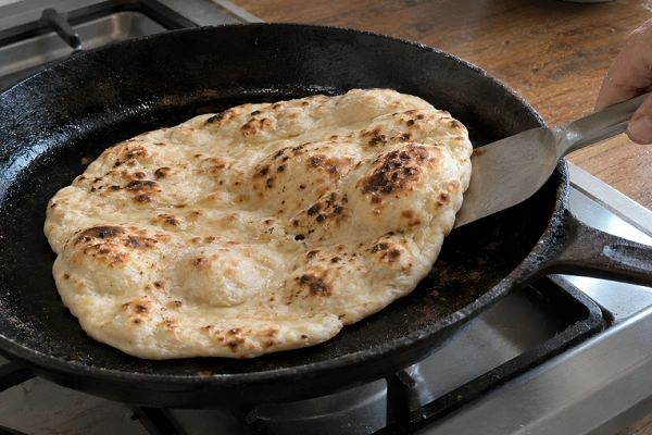 Naan bread bubbling and charring inside a hot black cast iron skillet on a gas stove