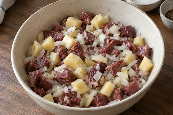 A white ceramic bowl filled with finely diced raw beef, potatoes, and chopped onions seasoned with black pepper.