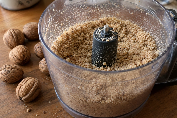 Finely ground walnuts inside a food processor bowl next to whole walnuts.