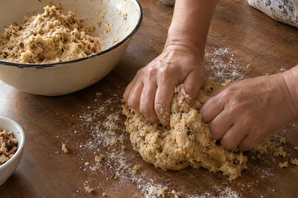 Hands kneading firm walnut-speckled dough on a flour-dusted wooden surface.