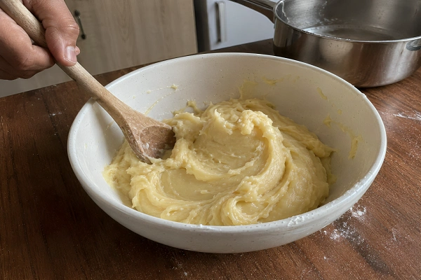 A person stirring smooth choux pastry dough in a white bowl with a wooden spoon