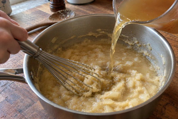 Pouring liquid broth into a pan while whisking continuously to make a lump-free gravy.
