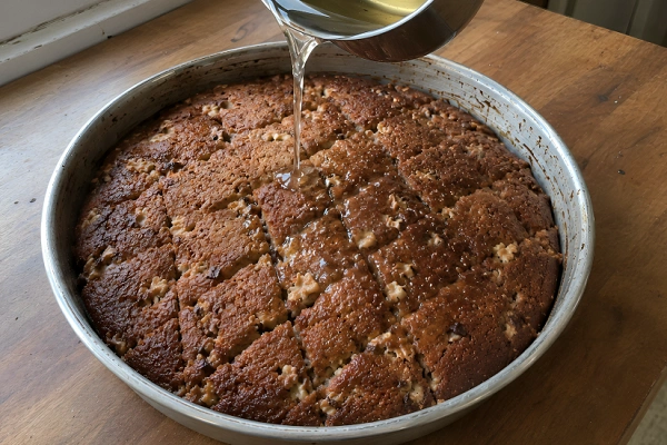 Pouring warm sugar syrup from a small pot over a sliced Shendetlie cake in a baking tray