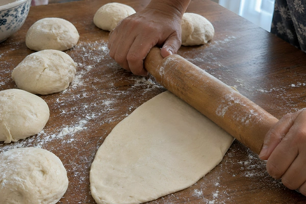 Rolling pin flattening a ball of dough into a classic oval naan shape next to shaped dough balls