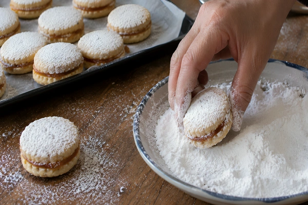 Hands coating a jam-filled Vanilice sandwich cookie in a bowl of vanilla powdered sugar.