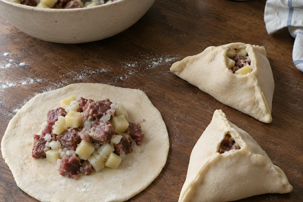 Raw beef and potato filling placed on a flat dough circle next to folded triangular pastries.