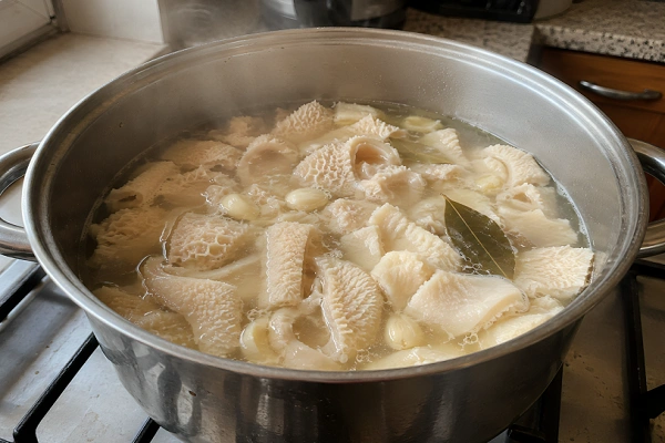 Whole cleaned beef tripe simmering with garlic cloves and bay leaves in a steel pot.
