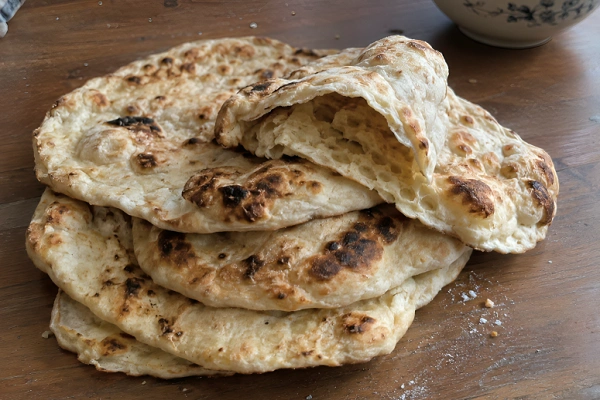 A stack of homemade naan breads with golden charred spots wrapped in a checkered towel