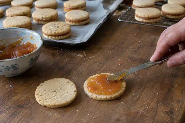 Spreading a layer of apricot jam onto a small baked cookie round with a butter knife.