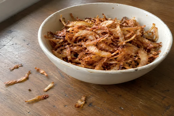 Thinly sliced onions fried to a deep golden brown crispness, piled high in a shallow ceramic dish.