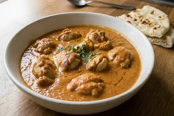 A white bowl filled with creamy butter chicken garnished with fresh coriander, served next to warm naan bread on a wooden table.